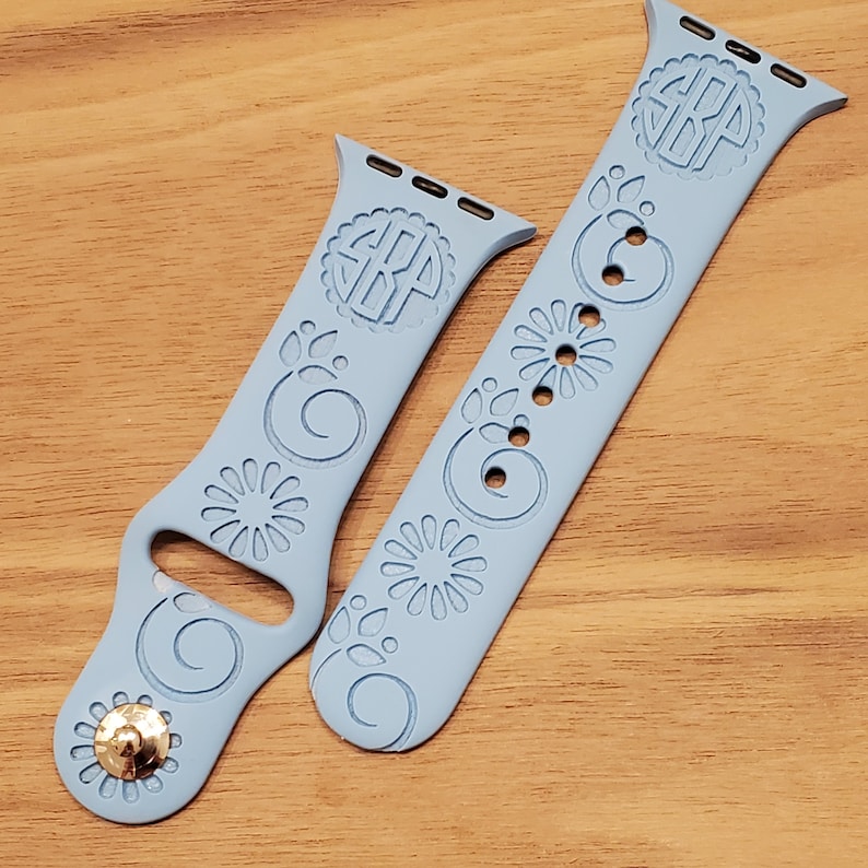 Pode incluir: Duas pulseiras Apple Watch azul claro com um design floral e um c&iacute;rculo com monograma. As pulseiras s&atilde;o de silicone e t&ecirc;m um acento dourado.