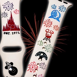 May include: Two white Apple Watch bands with colorful designs. One band features a red castle with the text "Est. 1971" and fireworks. The other band has a variety of colorful designs including fireworks, a castle, a tree, and a Mickey Mouse head.