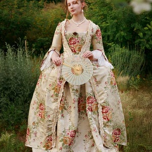Könnte beinhalten: Ein bodenlanges Kleid aus cremefarbenem Stoff mit einem roten und orangefarbenen Blumenmuster, langen Ärmeln mit weißen Spitzenbündchen, einem eckigen Ausschnitt und einer gerüschten Vorderkante. Dazu gehört ein runder, faltbarer Fächer.