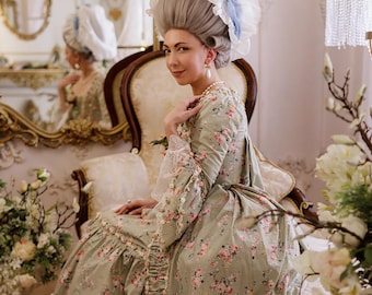 May include: A woman in a historical-style dress with a floral pattern, seated on a vintage chair. The dress is light green with pink flowers and lace details. She wears a large wig with a blue accent.