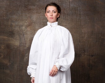 May include: A white, long-sleeved dress with a high, ruffled collar. The dress is made of a lightweight fabric and has a flowing silhouette. The model is wearing the dress with brown shoes.
