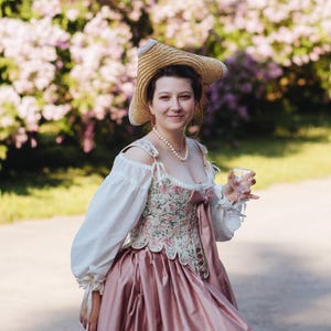 May include: A multi-piece outfit consisting of a straw hat with a wide brim, a white off-the-shoulder blouse with long sleeves, a floral print corset with a front ribbon tie, and a solid dusty pink floor-length skirt with a full silhouette.