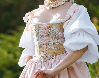 May include: A person in a historical costume, wearing a white bonnet with green trim, a cream-coloured lace choker, and a corset with a landscape design. The dress is light pink with white sleeves.