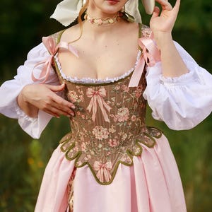 May include: A brown corset bodice featuring a floral pattern with pink bows, worn over a white blouse with puffed sleeves. The outfit includes a light pink skirt, a cream-colored wide-brimmed hat with ribbons, and a floral choker necklace.