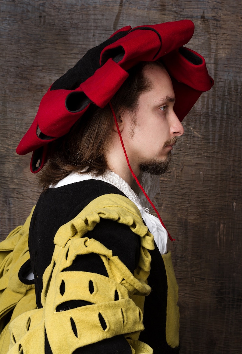 May include: A side profile of a person wearing a Renaissance-style costume. The outfit includes a black and red hat with a unique design, a black jacket with yellow accents, and a white collared shirt. The background is a textured wooden surface.