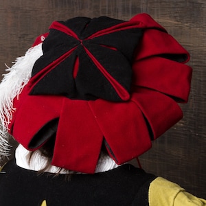 May include: A red and black hat with a white feather plume. The hat features a black top with a red trim and a series of red, fan-like panels. The hat is part of a costume.