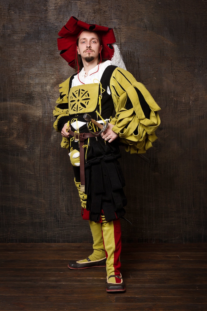 May include: A person in a Renaissance-style costume. The outfit features a large red hat, a yellow and black doublet, and black breeches. A sword hangs at the waist. The costume includes yellow and red leggings and black shoes. The background is a dark wood.