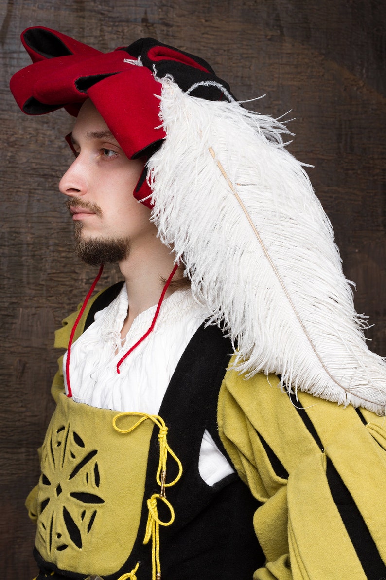 May include: A Renaissance-style costume featuring a red and black hat with a large white feather. The outfit includes a yellow and black vest with decorative cutouts and yellow ties, and a white shirt with a red neck cord.