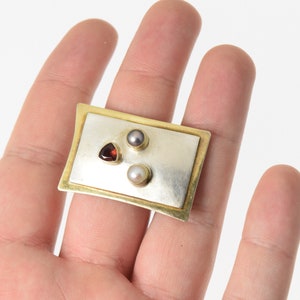 Pode incluir: Um broche retangular de prata e ouro com tr&ecirc;s pedras preciosas: uma pedra vermelha triangular, uma p&eacute;rola branca e uma p&eacute;rola cinza.