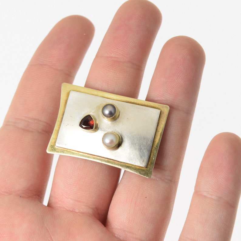 Pode incluir: Um broche retangular de prata e ouro com tr&ecirc;s pedras preciosas: uma pedra vermelha triangular, uma p&eacute;rola branca e uma p&eacute;rola cinza.