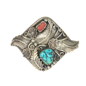 May include: A silver bracelet with a diamond-shaped design featuring turquoise and coral stones. The bracelet is decorated with intricate floral and feather motifs.