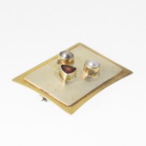 Pode incluir: Um broche retangular de ouro e prata com tr&ecirc;s pedras preciosas. O broche tem uma pedra preciosa triangular vermelha, uma p&eacute;rola branca e uma p&eacute;rola cinza.
