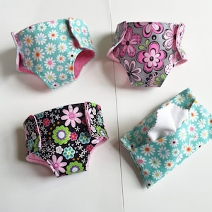 May include: Four handmade doll diapers with floral patterns. The diapers are made of fabric and have pink trim. There is also a small fabric pouch with a white tissue inside.