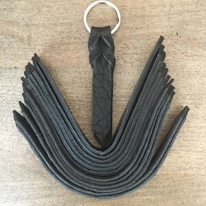 Puede incluir: Llavero de cuero negro con un lazo trenzado y un anillo de llave plateado. El llavero tiene un detalle de flecos.