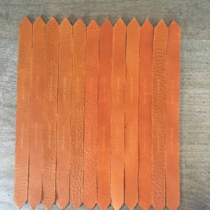 Puede incluir: Un conjunto de 15 tiras de cuero naranja con extremos puntiagudos, dispuestas en fila sobre una superficie de madera.