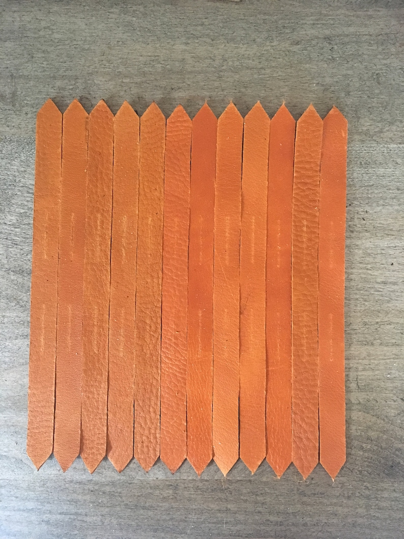 Puede incluir: Un conjunto de 15 tiras de cuero naranja con extremos puntiagudos, dispuestas en fila sobre una superficie de madera.