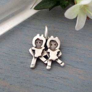 May include: Silver charm featuring two stylized figures of children holding hands. The charm is a simple, minimalist design.