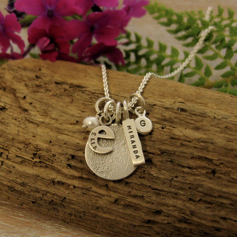 May include: Silver necklace with multiple charms, including a fingerprint charm with the letter "E" and the name "Ella" engraved, a charm with the letter "G", a charm with the name "Miranda" engraved, and a pearl bead.
