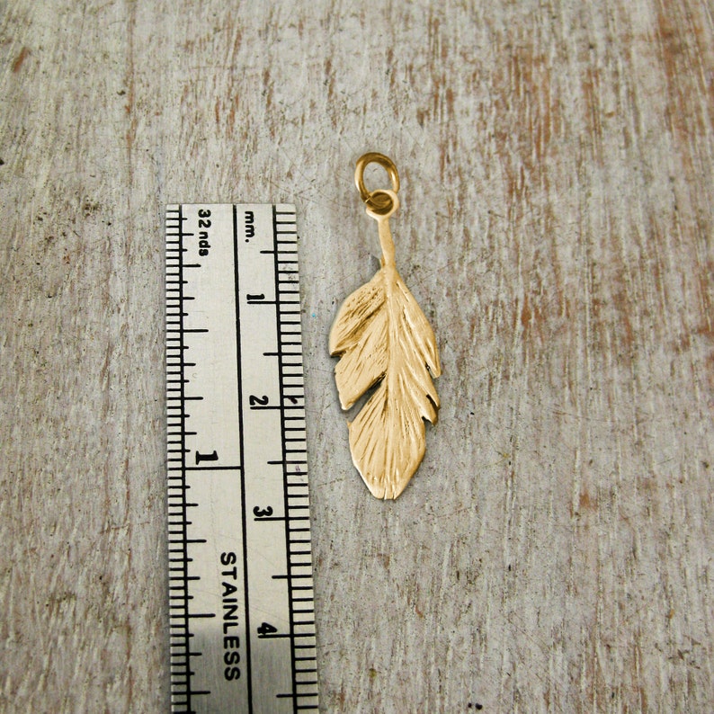 May include: A gold-colored feather charm pendant, approximately 3 cm long, is shown next to a ruler for size reference.