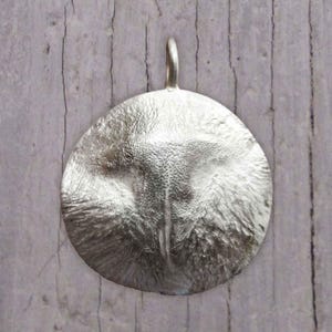 May include: A silver pendant in the shape of a circle with a textured surface. The pendant has a loop at the top for attaching a chain.