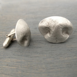 May include: A pair of silver cufflinks with a textured, organic shape. The cufflinks are made of metal and have a brushed finish.