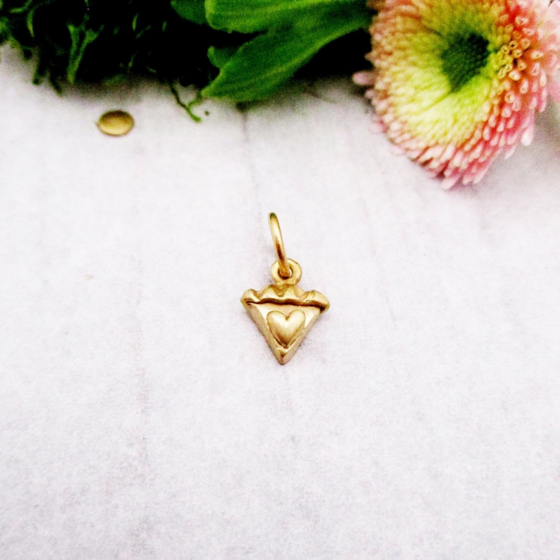 Puede incluir: Un peque&ntilde;o colgante dorado con un dise&ntilde;o en forma de coraz&oacute;n dentro de una forma triangular. El colgante tiene un lazo en la parte superior para sujetarlo a una cadena. El colgante es probablemente de metal y es una joya.