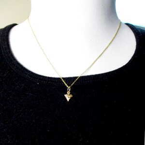 Puede incluir: Un delicado collar de oro con un peque&ntilde;o colgante en forma de diamante. El colgante cuelga de una fina cadena de oro. El collar se exhibe sobre un busto de maniqu&iacute; blanco, sobre un fondo negro.