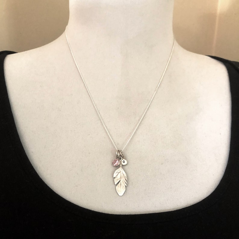 May include: A silver chain necklace with a silver feather charm, a small pink crystal charm, and a silver charm with the letter "G" on it.