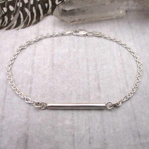 May include: A delicate silver bracelet featuring a bar pendant. The bracelet has a chain-link design and a secure clasp. The bar pendant is centered on the bracelet. The jewelry is displayed on a light gray surface.