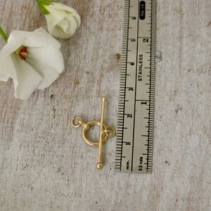 May include: A gold-toned metal toggle clasp with a round ring and a bar. The clasp is measured against a ruler, showing it is approximately 1 inch long.