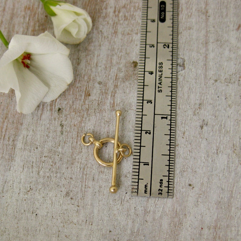 May include: A gold-toned metal toggle clasp with a round ring and a bar. The clasp is measured against a ruler, showing it is approximately 1 inch long.