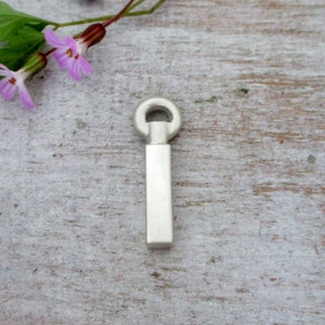 May include: A silver pendant with a rectangular body and a circular loop at the top. The pendant is displayed on a light-coloured wooden surface, with delicate purple flowers and green stems in the background.