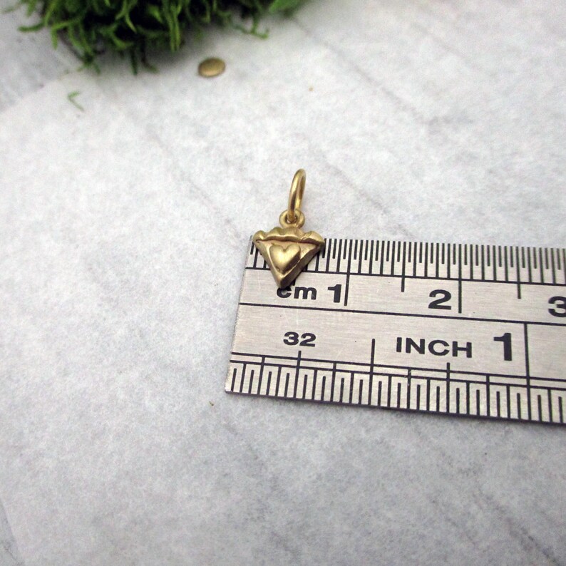 Puede incluir: Un peque&ntilde;o colgante dorado con un coraz&oacute;n en el centro y un dise&ntilde;o triangular. El colgante mide aproximadamente 1,3 cm de ancho y se muestra junto a una regla para la escala. El colgante tiene un peque&ntilde;o lazo en la parte superior para una cadena.