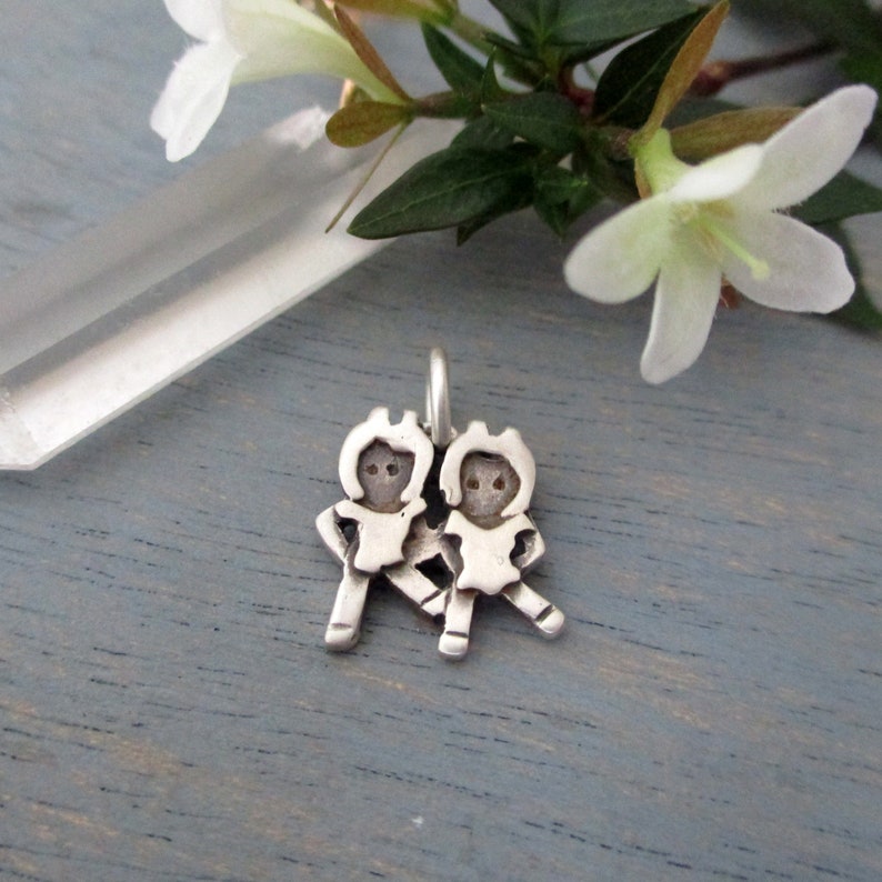 May include: Silver charm featuring two stylized figures of children holding hands. The charm is a simple, minimalist design.