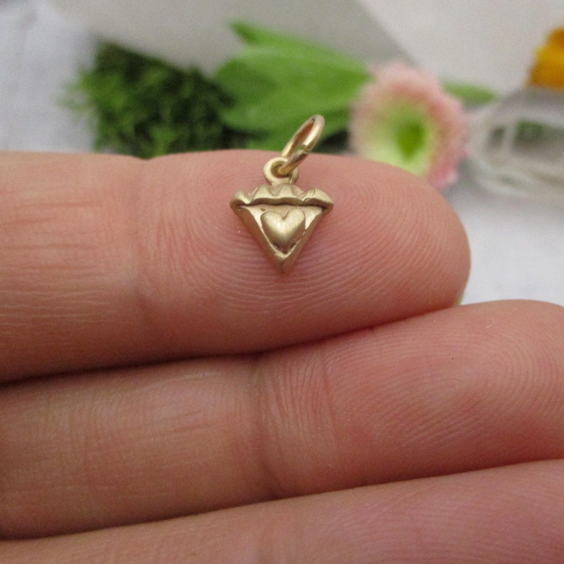 Puede incluir: Un peque&ntilde;o dije dorado con un coraz&oacute;n en el centro y una parte superior en forma de corona. El dije tiene forma triangular y tiene un lazo en la parte superior para sujetarlo a un collar o pulsera. El dije se sostiene entre dos dedos.