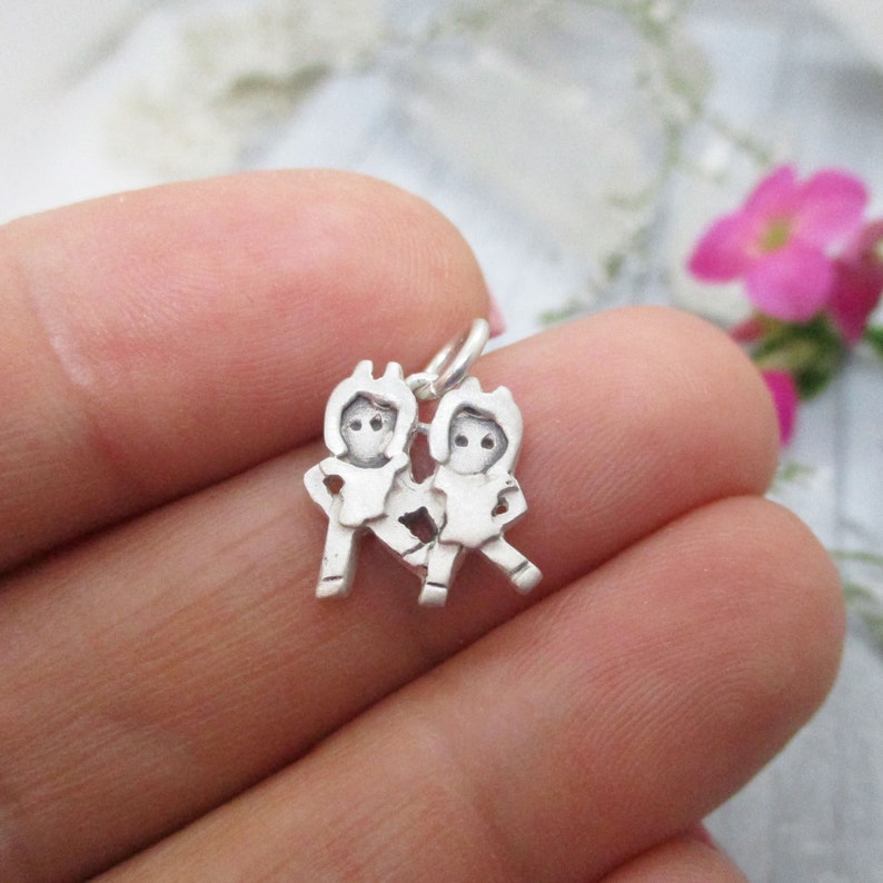 May include: Silver charm featuring two stylized figures of people holding hands. The charm is designed to be added to a necklace or bracelet.