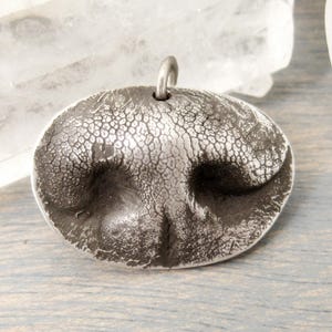 May include: A silver-toned pendant shaped like a dog's nose, featuring a textured surface and a small loop at the top for a chain. The pendant has a detailed, realistic design, perfect for animal lovers.