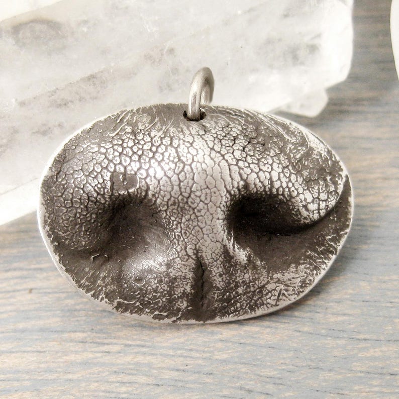May include: A silver-toned pendant shaped like a dog's nose, featuring a textured surface and a small loop at the top for a chain. The pendant has a detailed, realistic design, perfect for animal lovers.