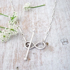 May include: A silver necklace featuring an infinity symbol charm with a small silver rod passing through its centre.