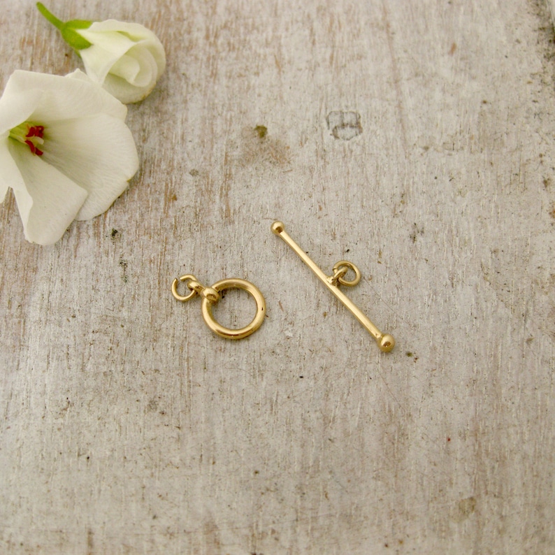 May include: Gold-toned metal toggle clasp with a round ring and a straight bar. The clasp is on a light wood surface.