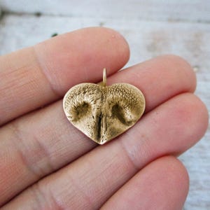 May include: A heart-shaped pendant with a textured surface, featuring a detailed impression of a dog's nose. The pendant is a warm gold color and has a small loop at the top for a chain. The pendant is held in a hand.