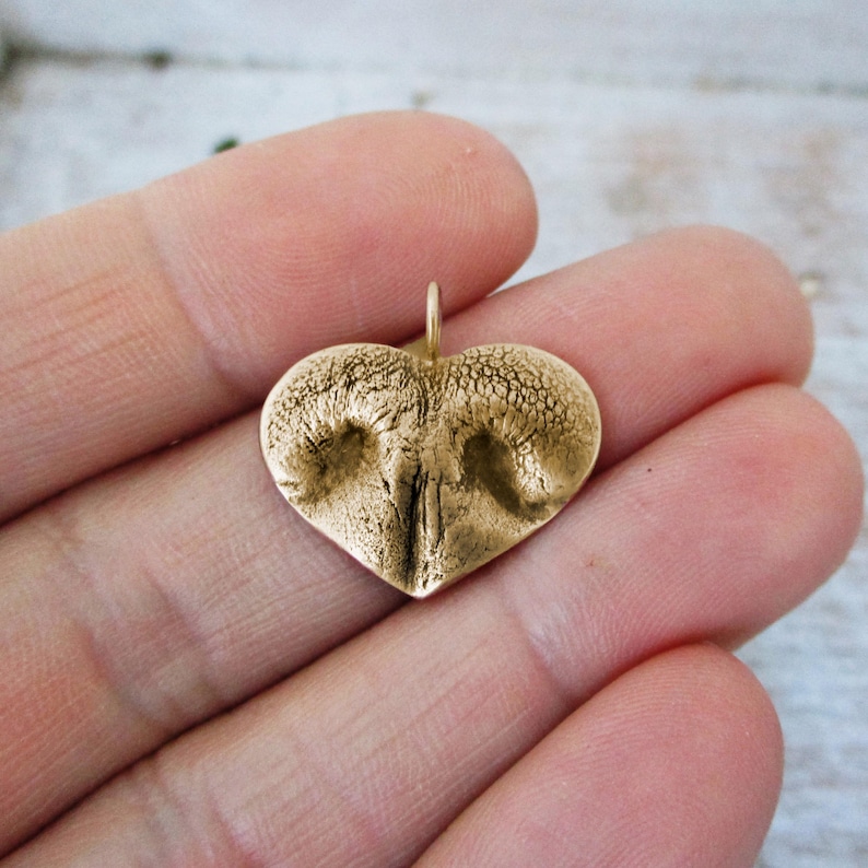 May include: A heart-shaped pendant with a textured surface, featuring a detailed impression of a dog's nose. The pendant is a warm gold color and has a small loop at the top for a chain. The pendant is held in a hand.