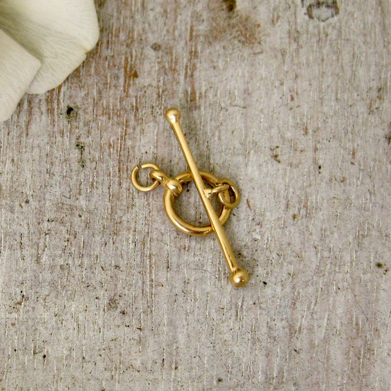 May include: Gold-toned metal toggle clasp with a round ring and a straight bar. The clasp is designed for necklaces, bracelets, or other jewelry.