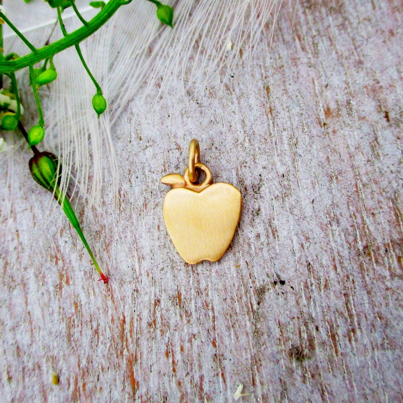 May include: A gold-colored apple charm pendant. The charm is a simple, solid design with a smooth, shiny finish.