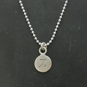 Puede incluir: Un collar de plata con un colgante redondo que presenta el s&iacute;mbolo del zod&iacute;aco de Capricornio.