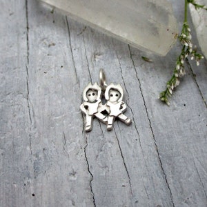 May include: Silver charm featuring two stylized figures of children holding hands. The charm is on a wooden surface.