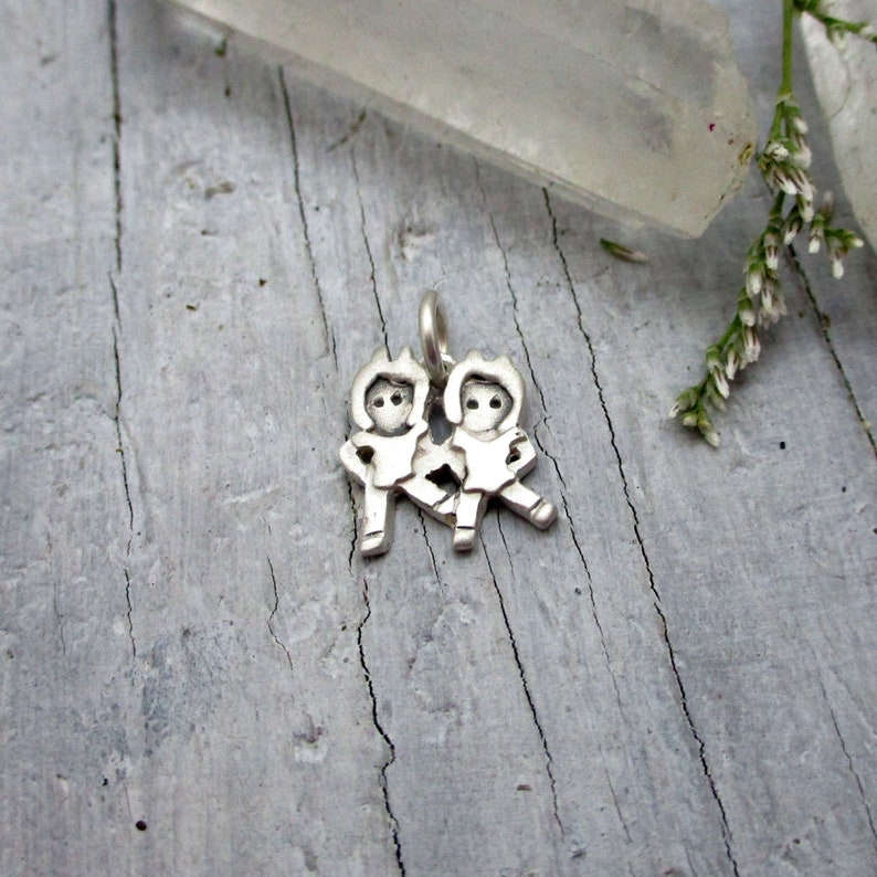 May include: Silver charm featuring two stylized figures of children holding hands. The charm is on a wooden surface.