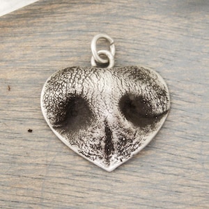 May include: A heart-shaped pendant with a textured, silver finish. The pendant features a detailed impression of a dog's nose, with dark areas representing the nostrils. A small loop is at the top for a chain.