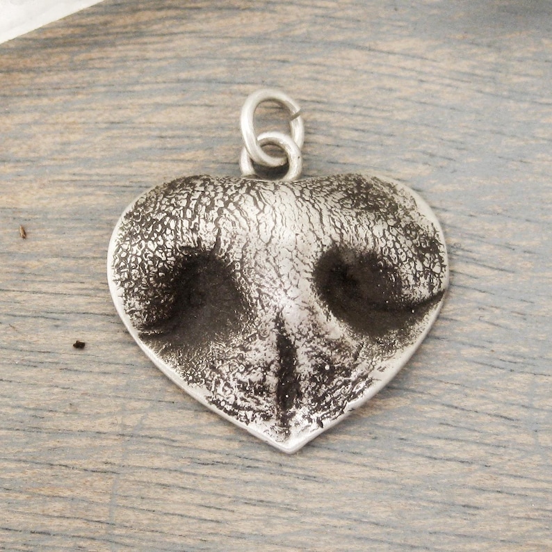 May include: A heart-shaped pendant with a textured, silver finish. The pendant features a detailed impression of a dog's nose, with dark areas representing the nostrils. A small loop is at the top for a chain.