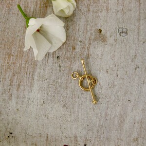 May include: Gold-toned metal toggle clasp with a round ring and a straight bar. The clasp is on a light wood surface.